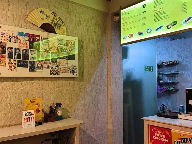Close-up of the Wang House Korean Theme Cafe counter, featuring a wall collage of K-Pop photos (BTS), a traditional fan, and an illuminated menu board.