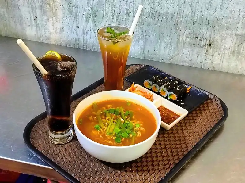 Korean Theme Cafe meal with spicy ramen, kimbap, kimchi, dipping sauce and chilled iced drinks served on tray