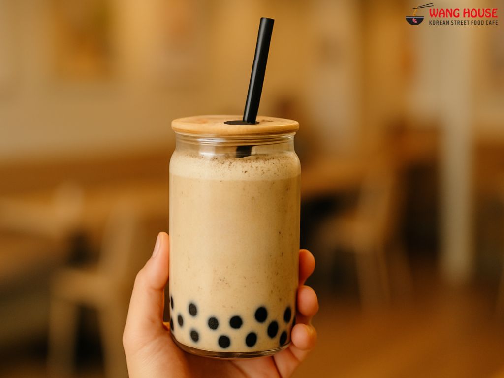 trawberries and Cream boba tea in Delhi from The Wang House, Laxmi Nagar – A pastel pink milk tea served in a clear glass jar with a golden lid and black straw, topped with a light froth and filled with black tapioca pearls. The drink radiates a sweet, fruity aroma and creamy richness, set against a stylish background, capturing the essence of a refreshing Korean-style boba tea in East Delhi.