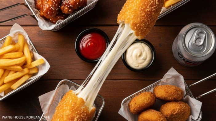 Regional Korean Food from Wang House Korean Cafe. An overhead shot of a Korean cheese corn dog being pulled apart, creating a long, gooey cheese stretch. The corn dog is surrounded by other popular street foods, including a basket of Korean Fried Chicken, a tray of golden french fries, and a bowl of fried chicken nuggets. Two small dipping bowls of ketchup and white sauce are also visible on the dark wooden table, alongside a can of soda.