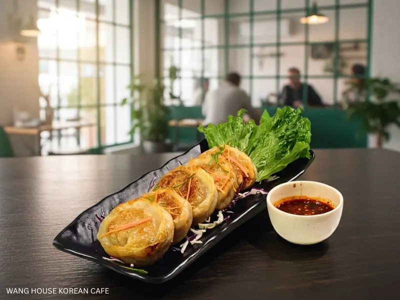 Best Korean Cafe: A platter of golden-brown pan-fried dumplings (mandu) served with fresh lettuce and a spicy dipping sauce, in a stylish cafe with large windows. Wang House Korean Cafe.
