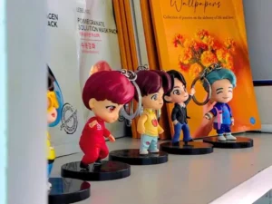 Four miniature K-pop-style chibi figurines with brightly colored hair (red, yellow, black, and turquoise) stand on a white shelf in a Korean Theme Cafe. They are posed in a line next to Korean face mask packaging, reflecting the cafe's blend of K-pop and Korean culture.