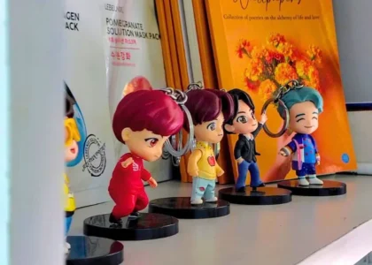 Four miniature K-pop-style chibi figurines with brightly colored hair (red, yellow, black, and turquoise) stand on a white shelf in a Korean Theme Cafe. They are posed in a line next to Korean face mask packaging, reflecting the cafe's blend of K-pop and Korean culture.