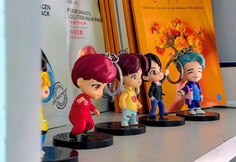 Four miniature K-pop-style chibi figurines with brightly colored hair (red, yellow, black, and turquoise) stand on a white shelf in a Korean Theme Cafe. They are posed in a line next to Korean face mask packaging, reflecting the cafe's blend of K-pop and Korean culture.